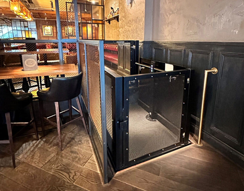 Compact, industrial-style platform lift with black mesh enclosure in a restaurant setting.