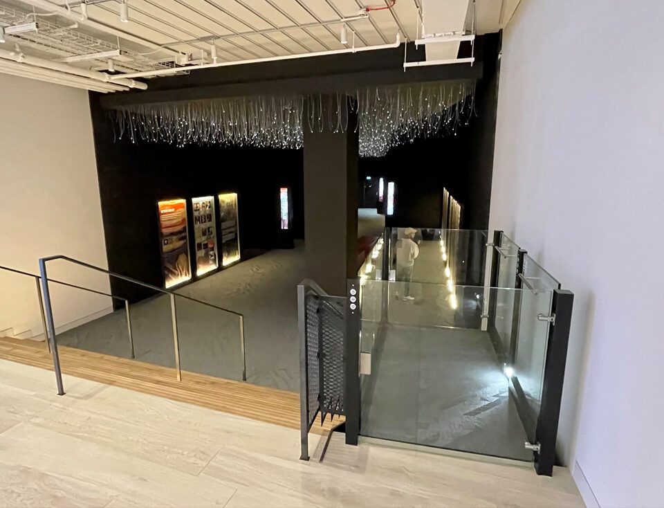 Stylish platform lift with glass enclosure descending into a modern, dark exhibition space.